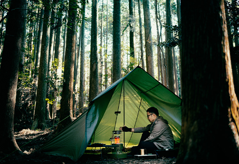 Calentador redondo de parafina (keroseno) Toyotomi GEAR MISSION en una tienda de campaña en medio de un bosque, calentando a una persona dentro de la tienda.