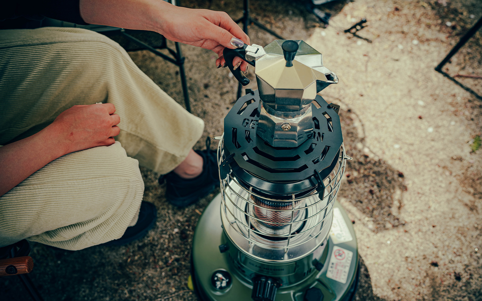 Toyotomi GEAR MISSION round heater in use by a person at a campsite, providing warmth outdoors while also heating up her coffee.
